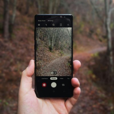 A hand holds a black smartphone showing the camera app interface with a photo preview of a leafy forest path. The background is a blurred forest.