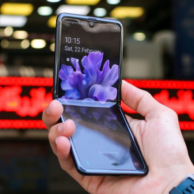 A hand holding a Samsung Galaxy Z Flip phone partially folded, showing the lock screen with a digital clock and a floral background.
